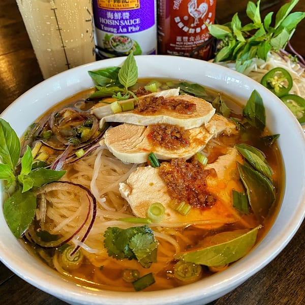 Chicken pho with basil, red onions, and chili paste, with hoisin and sriracha bottles in the background.