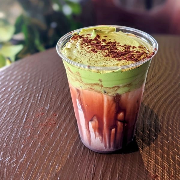 Presotea drink with green matcha foam and red liquid, topped with cocoa powder, on a wooden table.