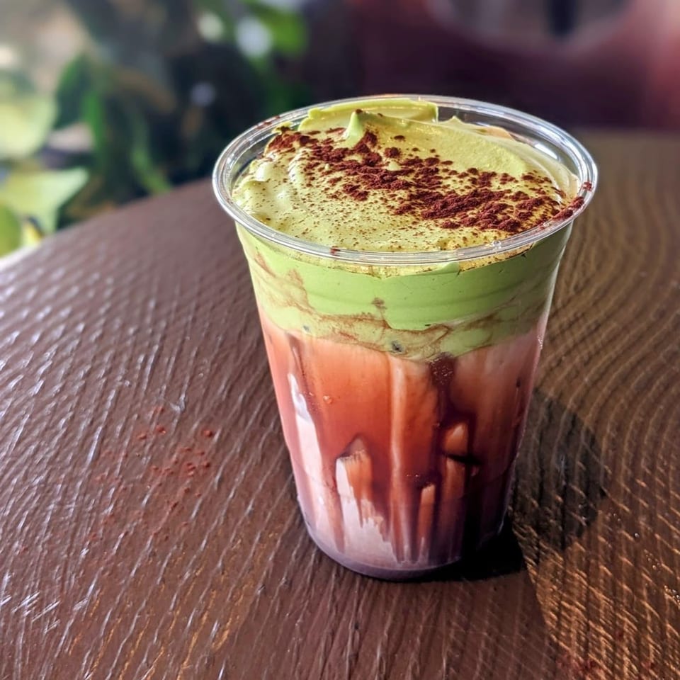 Presotea drink with green matcha foam and red liquid, topped with cocoa powder, on a wooden table.