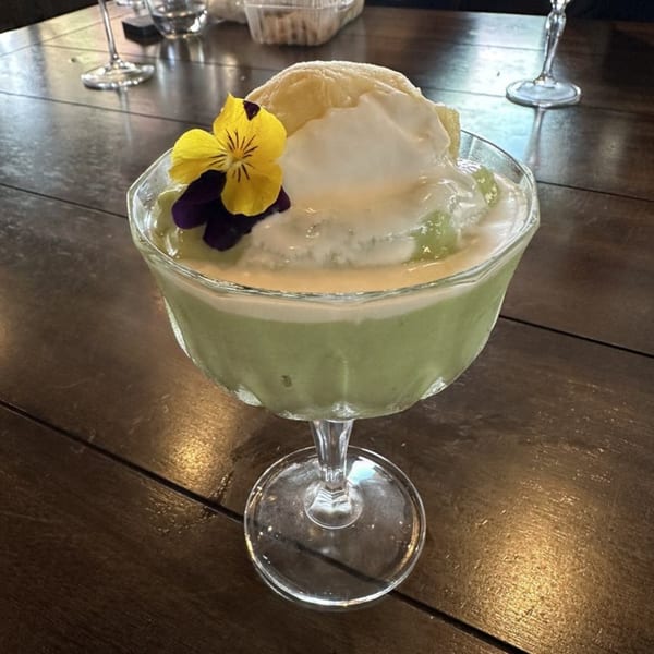 Green Vietnamese dessert with white cream, a yellow flower, and purple petals in a glass.